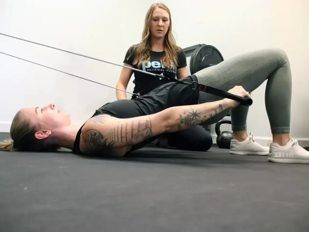 A woman with tattoos performs a glute bridge using resistance bands on a gym mat, guided by a trainer in athletic wear. The scene conveys focused physical training.