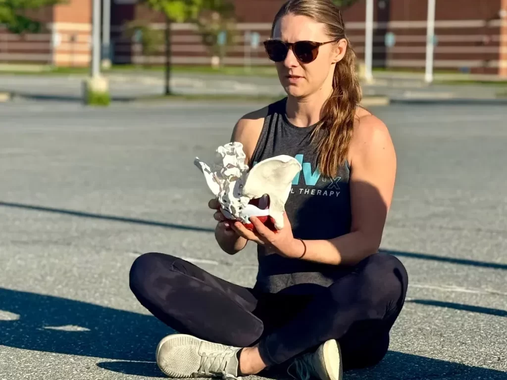 A woman sits cross-legged on asphalt, wearing sunglasses and holding a pelvic bone model. She appears focused, conveying a sense of calmness and study.
