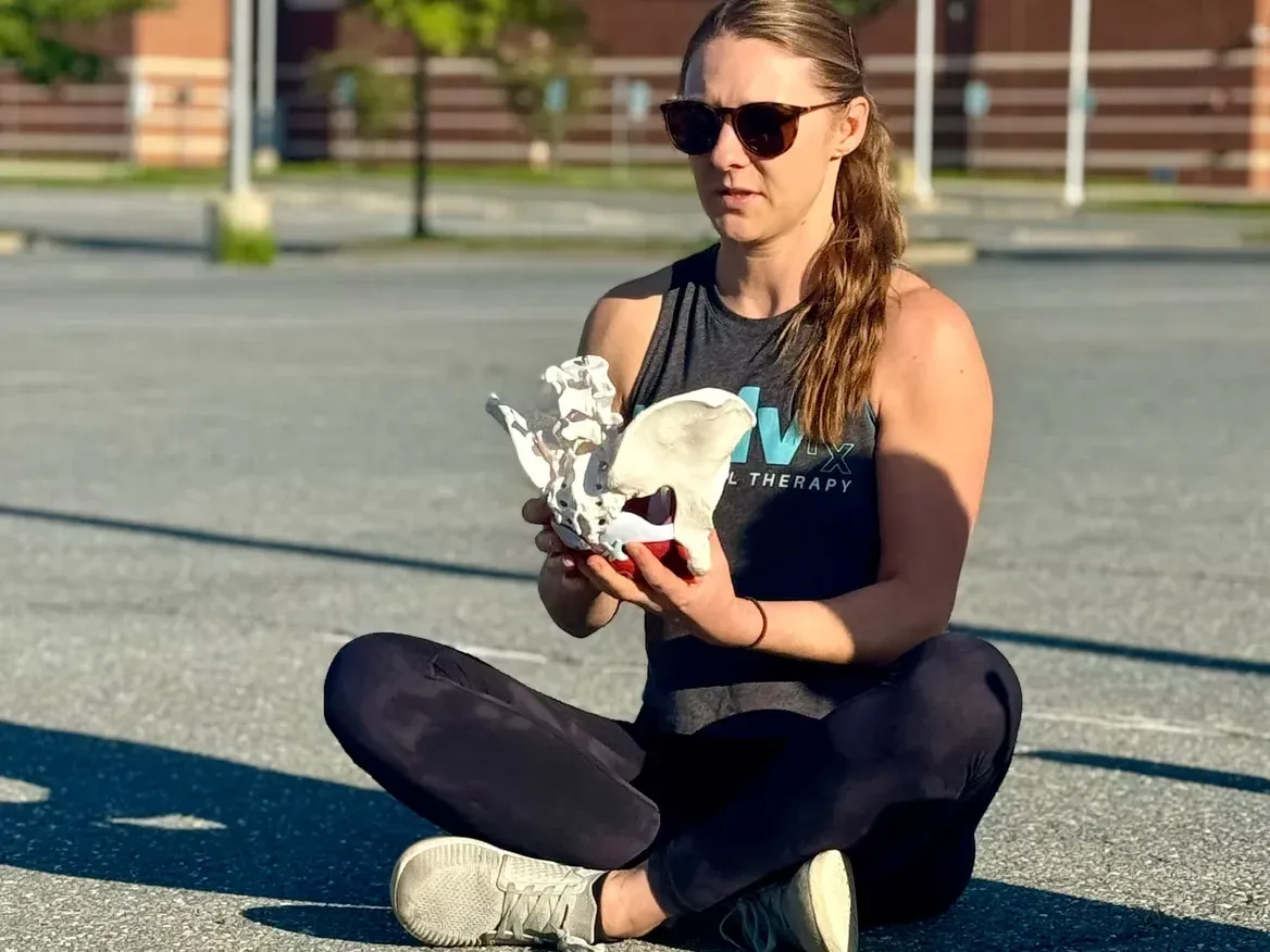 A woman sits cross-legged on asphalt, wearing sunglasses and holding a pelvic bone model. She appears focused, conveying a sense of calmness and study.