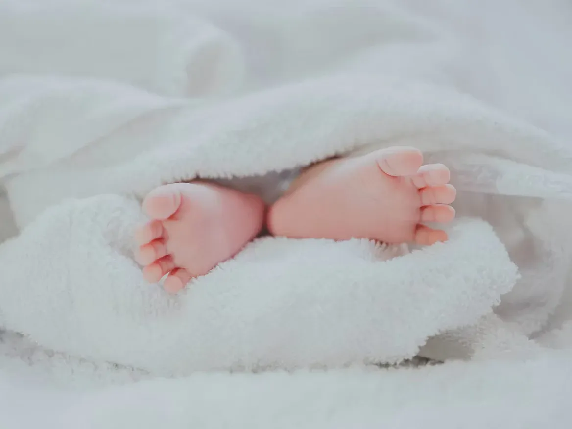 Tiny baby feet peek out from underneath a soft, white blanket. The scene is peaceful and cozy, conveying warmth and innocence.