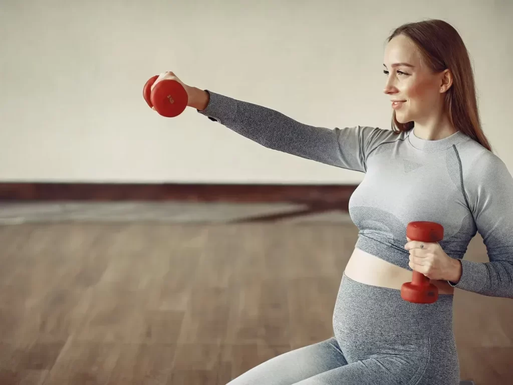 Pregnant woman in gray athletic wear exercises with red dumbbells in a serene studio setting. She smiles, embodying health and vitality.